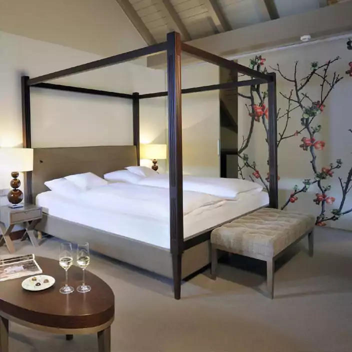 Helles Hotelzimmer mit Himmelbett, dekorativer Tapete und zwei Gläsern Weißwein auf dem Tisch